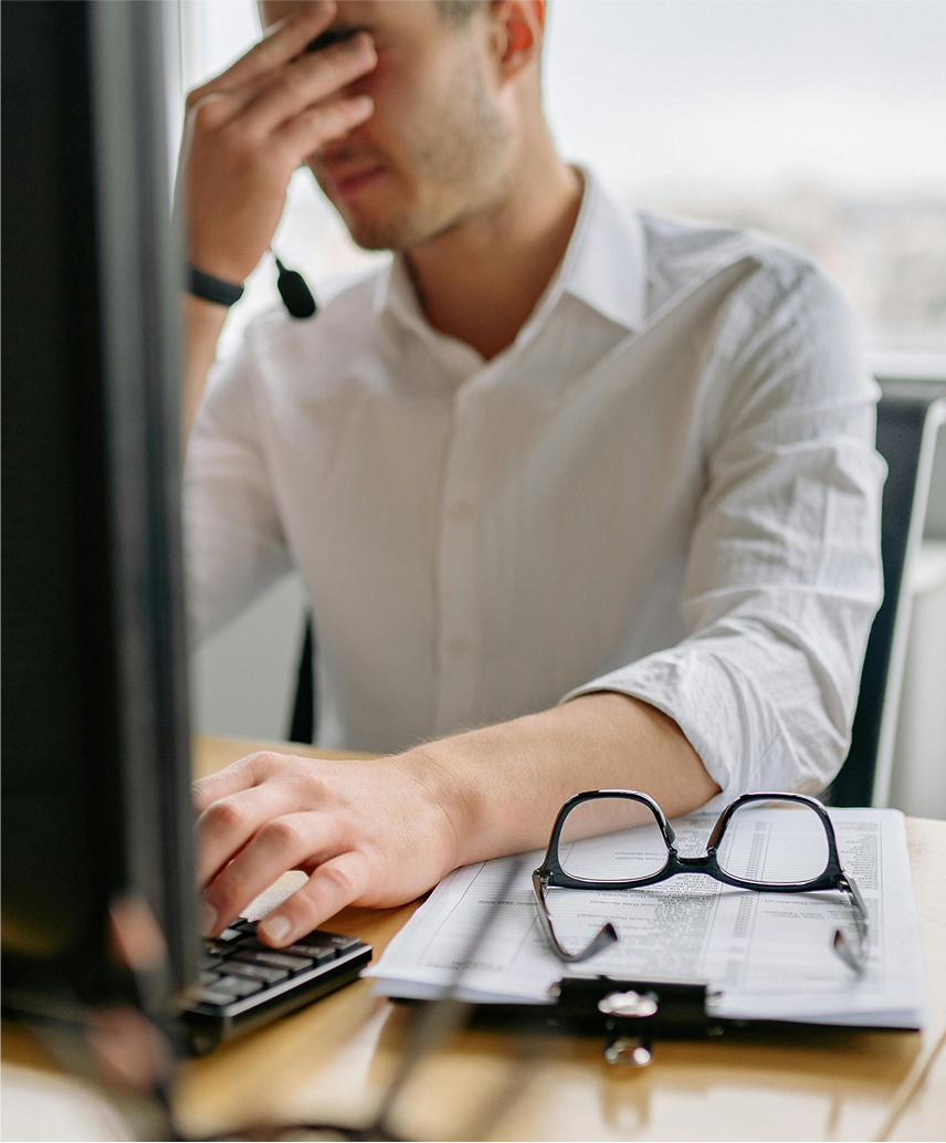 man sitting at computer looking stressed with his hand rubbing his eyes